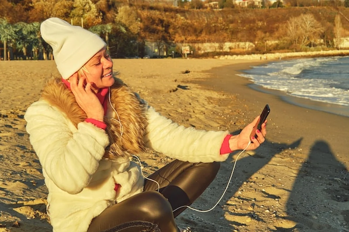 Kobieta w futrze i kapeluszu na plaży słuchając muzyki na smartfonie
